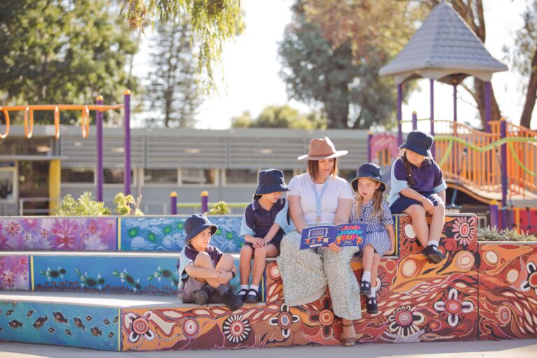 A Primary school teacher and children read a book in the shade outside.